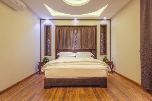 A bedroom with a large bed, wooden flooring, a ceiling light fixture, and a window with curtains.
