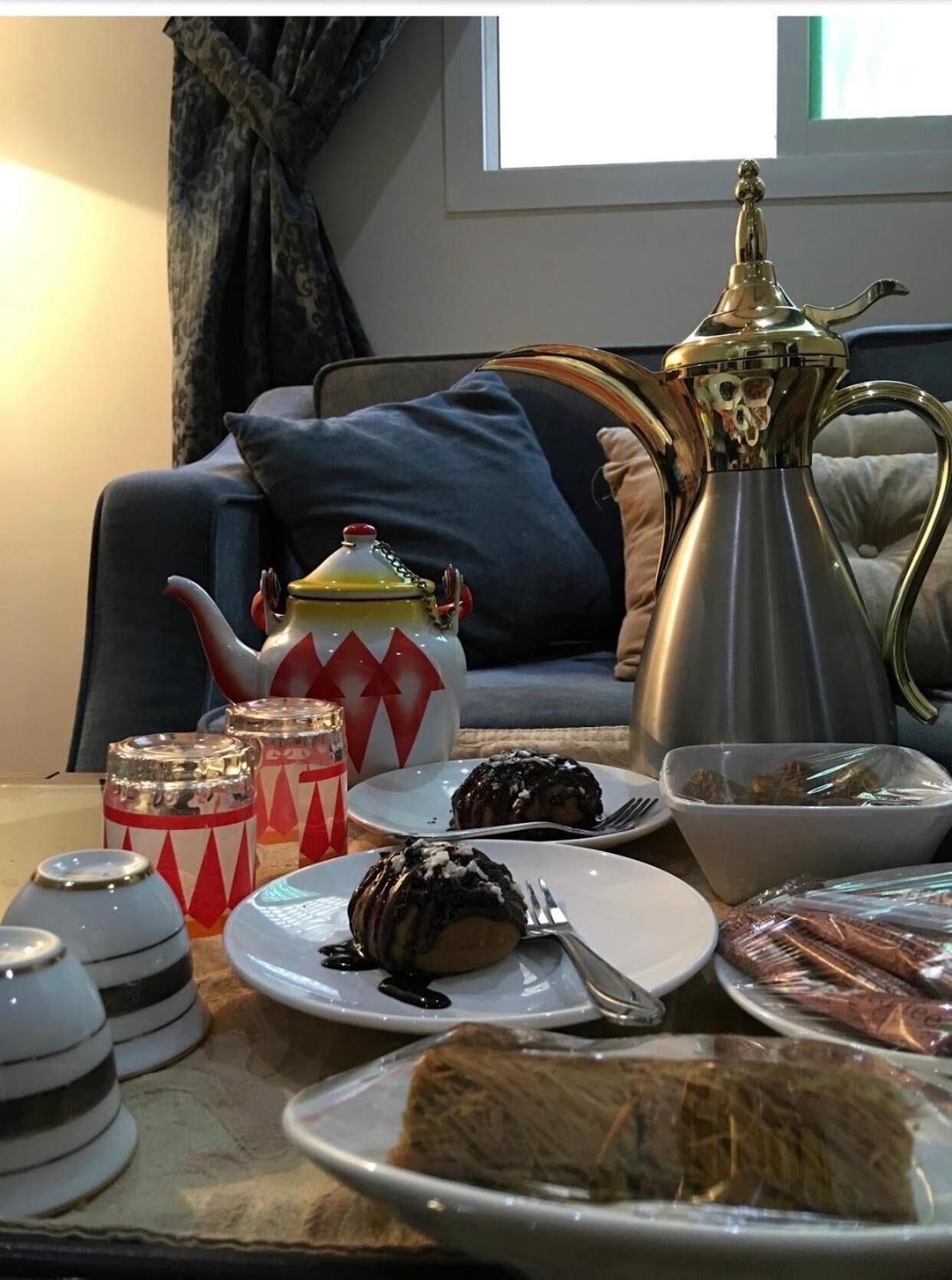A dining setup with a teapot, plates of food, and decorative jars on a table in a room with a sofa and curtains.