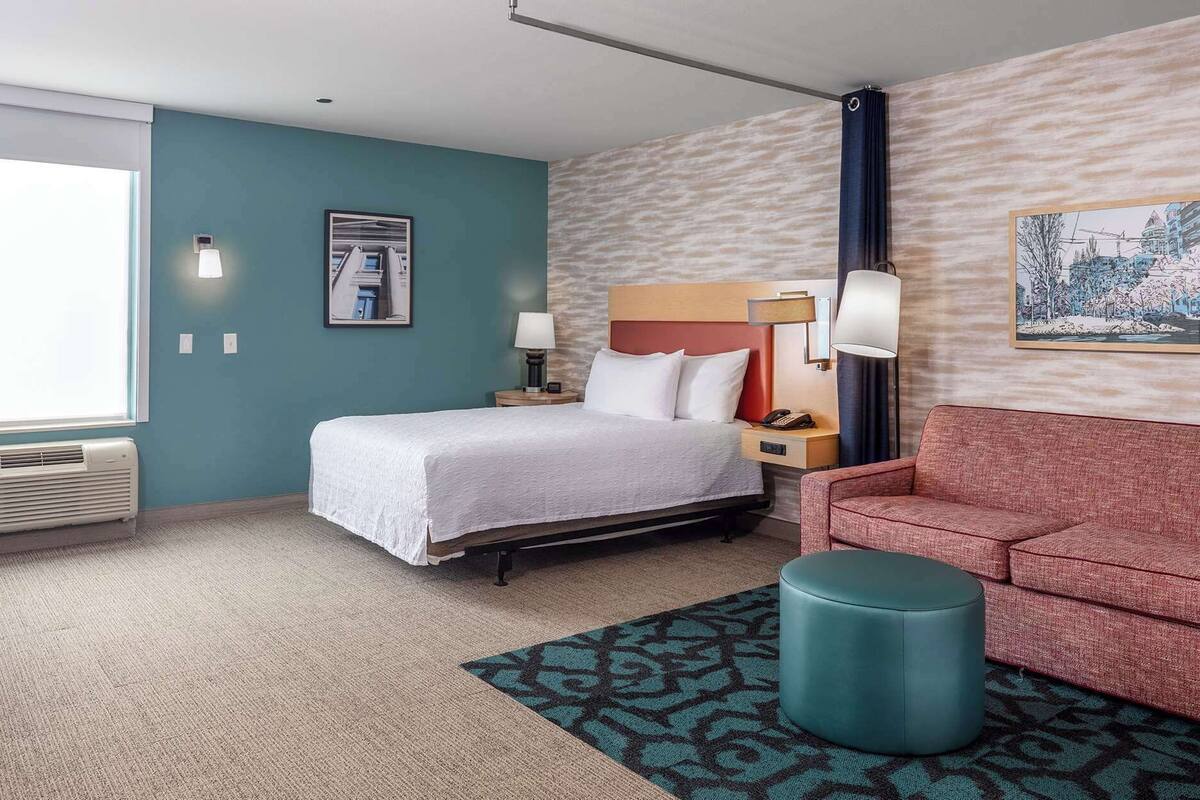 A hotel room with a bed, a red sofa, a teal ottoman, and a patterned rug.