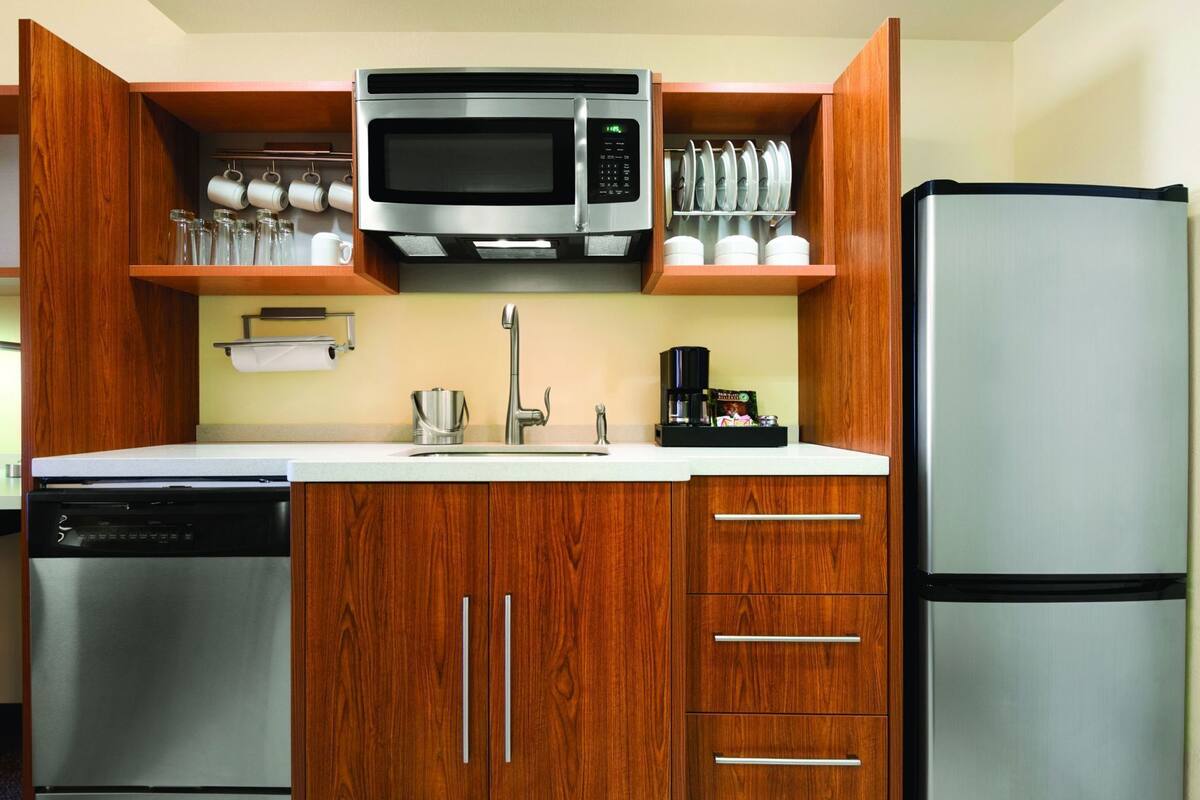 A compact kitchen with wooden cabinets, a stainless steel dishwasher, a microwave, a refrigerator, and a coffee maker.