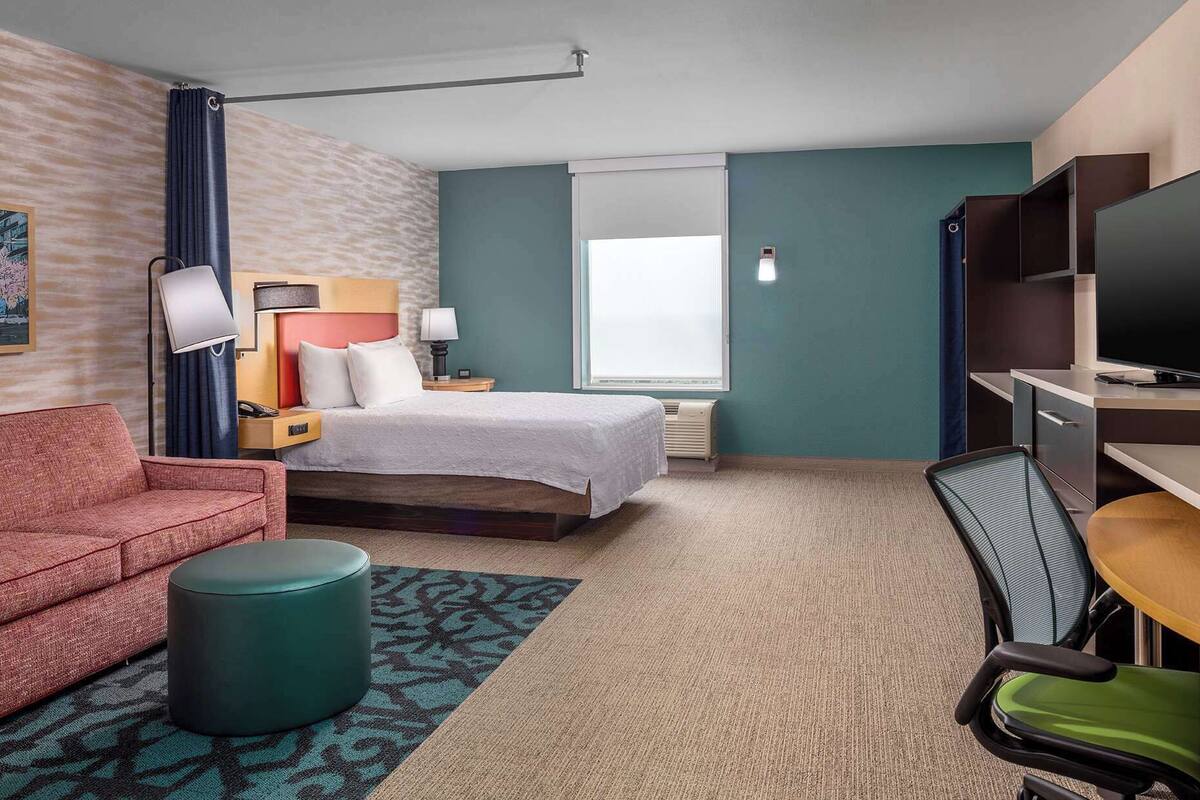 A hotel room with a bed, a desk with a TV, a sofa, a chair, and a patterned rug.