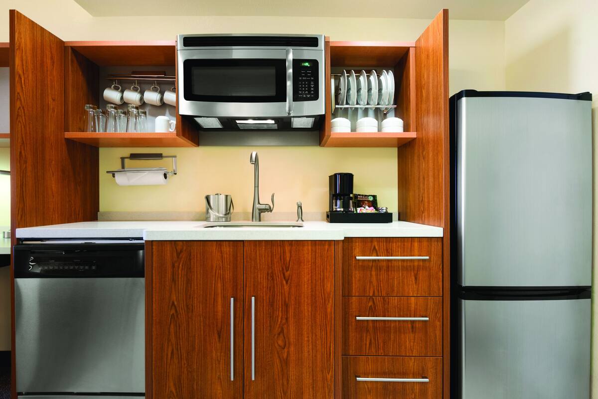 A compact kitchen with wooden cabinets, a stainless steel dishwasher, a microwave, a refrigerator, and a coffee maker.