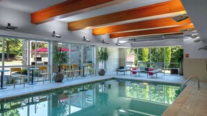 Une piscine intérieure aux parois en verre transparent, avec des poutres en bois au plafond et des espaces extérieurs aménagés avec des chaises et des parasols.
