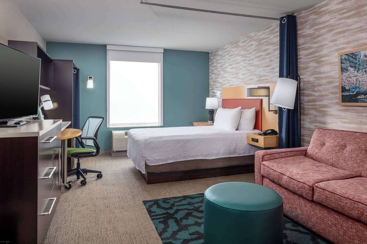 A hotel room with a bed, a desk with a chair, a sofa, a small ottoman, and a painting on the wall.