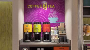 A coffee and tea station with three dispensers labeled 'Gourmet', 'Decaf', and 'Hot H2O'. There are trays of cups and a trash bin below.