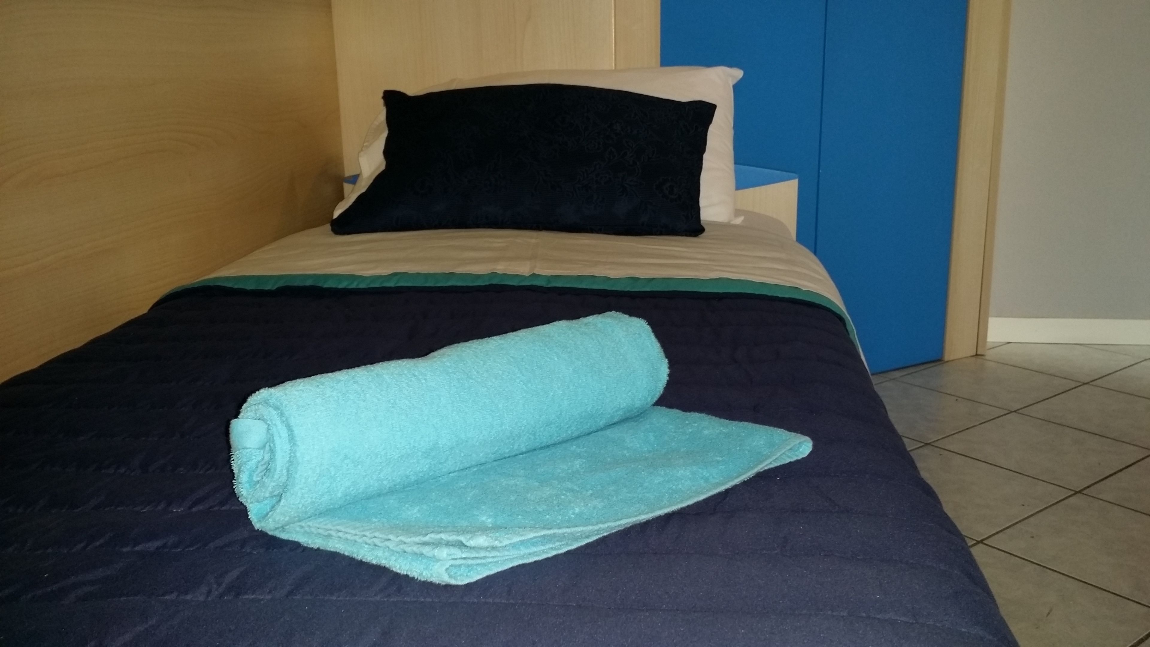A bed with a dark blue bedspread, a teal towel, and two pillows (one black and one white).