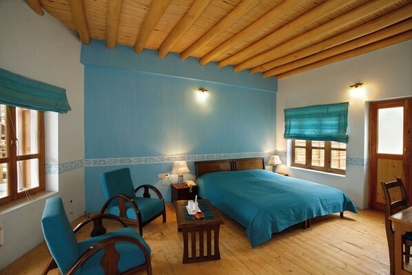 A room with a wooden ceiling, a bed with a blue bedspread, two chairs, a small table, and a window with a wooden shutter.