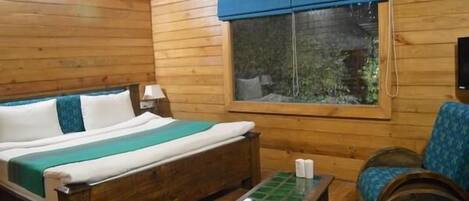 A wooden cabin room with a bed, a small table, a chair, and a window with a view of trees.
