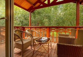 Área de estar coberta ao ar livre com móveis de vime e uma mesa de vidro, cercada por um deck de madeira e uma vista de folhagem densa.
