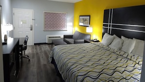 A hotel room with a bed, a desk with a chair, a sofa, and a bedside table with a lamp.