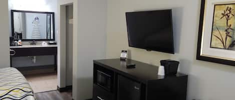 Una habitación de hotel con cama, escritorio, silla, televisión, microondas y un refrigerador pequeño.
