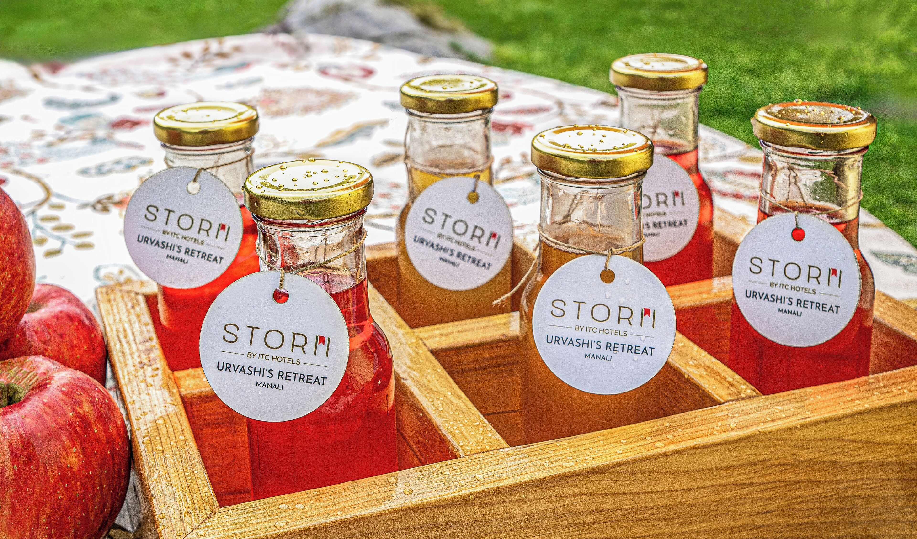 A wooden tray with several glass bottles labeled 'STORM URVASHI'S RETREAT MANALI' and a few apples.