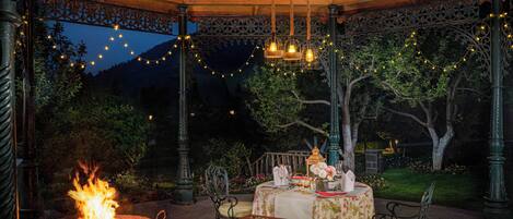 An outdoor gazebo with a fire pit, tables set for dining, and string lights.