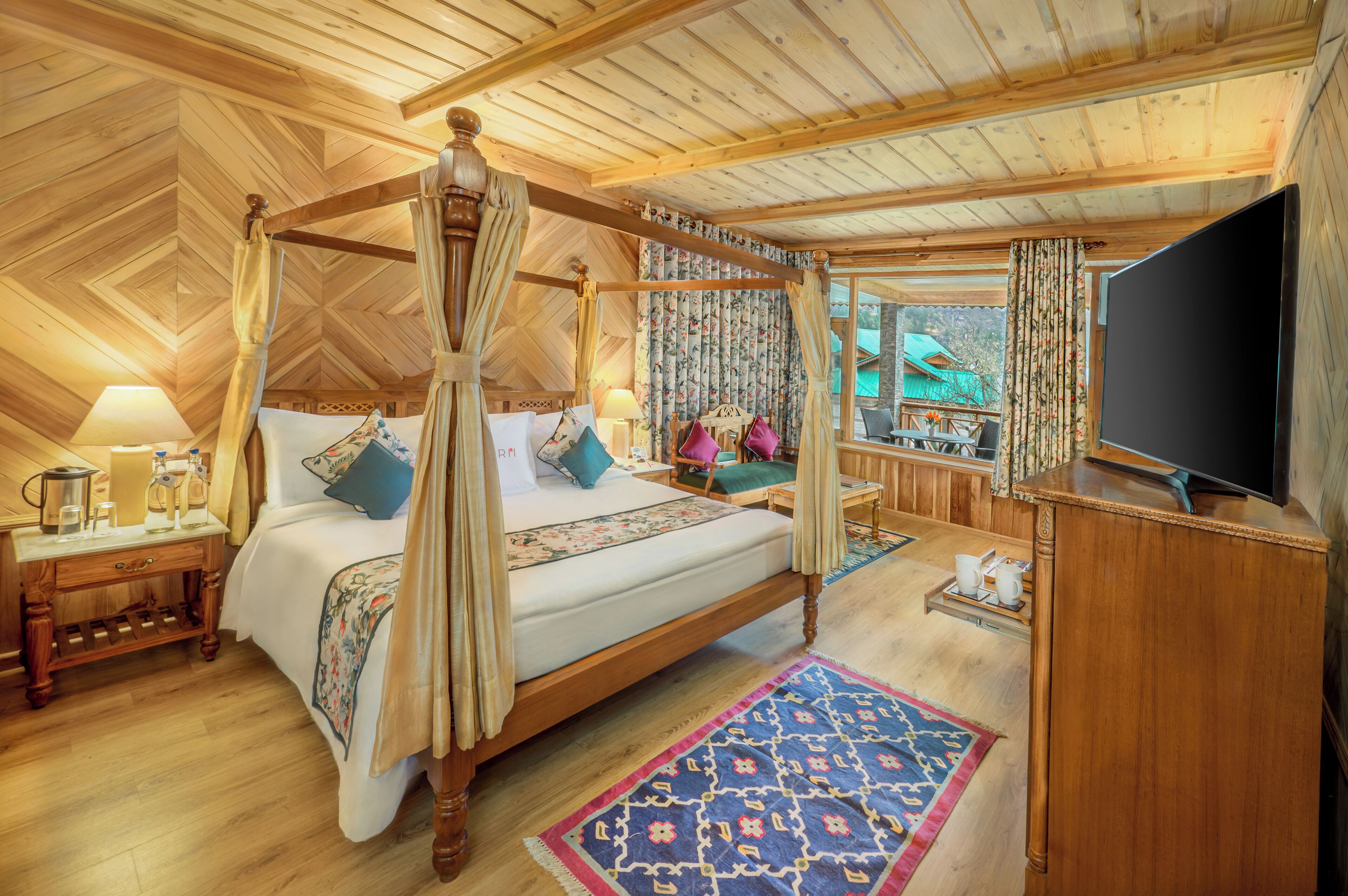 A four-poster bed with a canopy, a television on a wooden stand, a wooden nightstand with a lamp, and a view of the outdoors through a window.