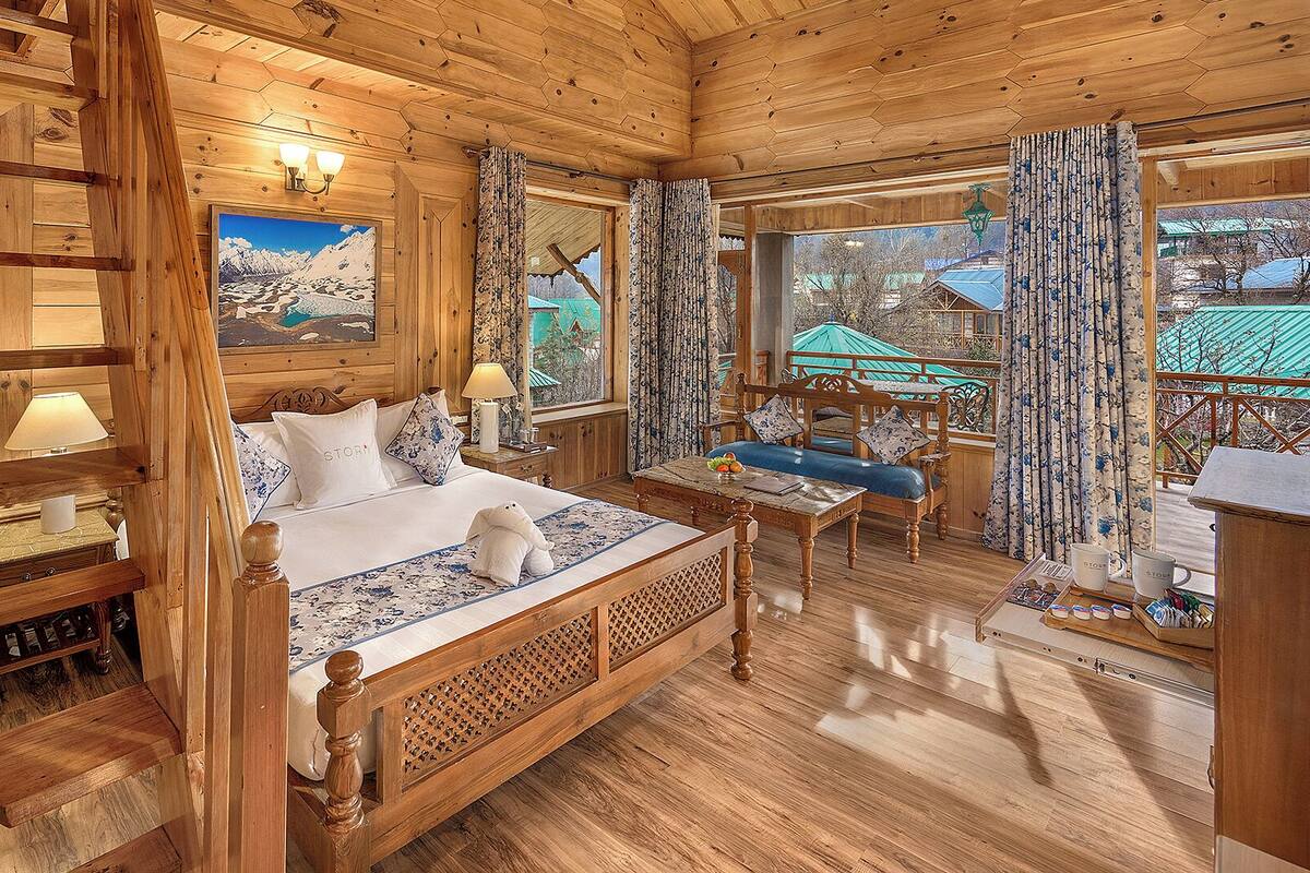 A wooden cabin room with a bed, a small table, and a view of a mountain landscape.