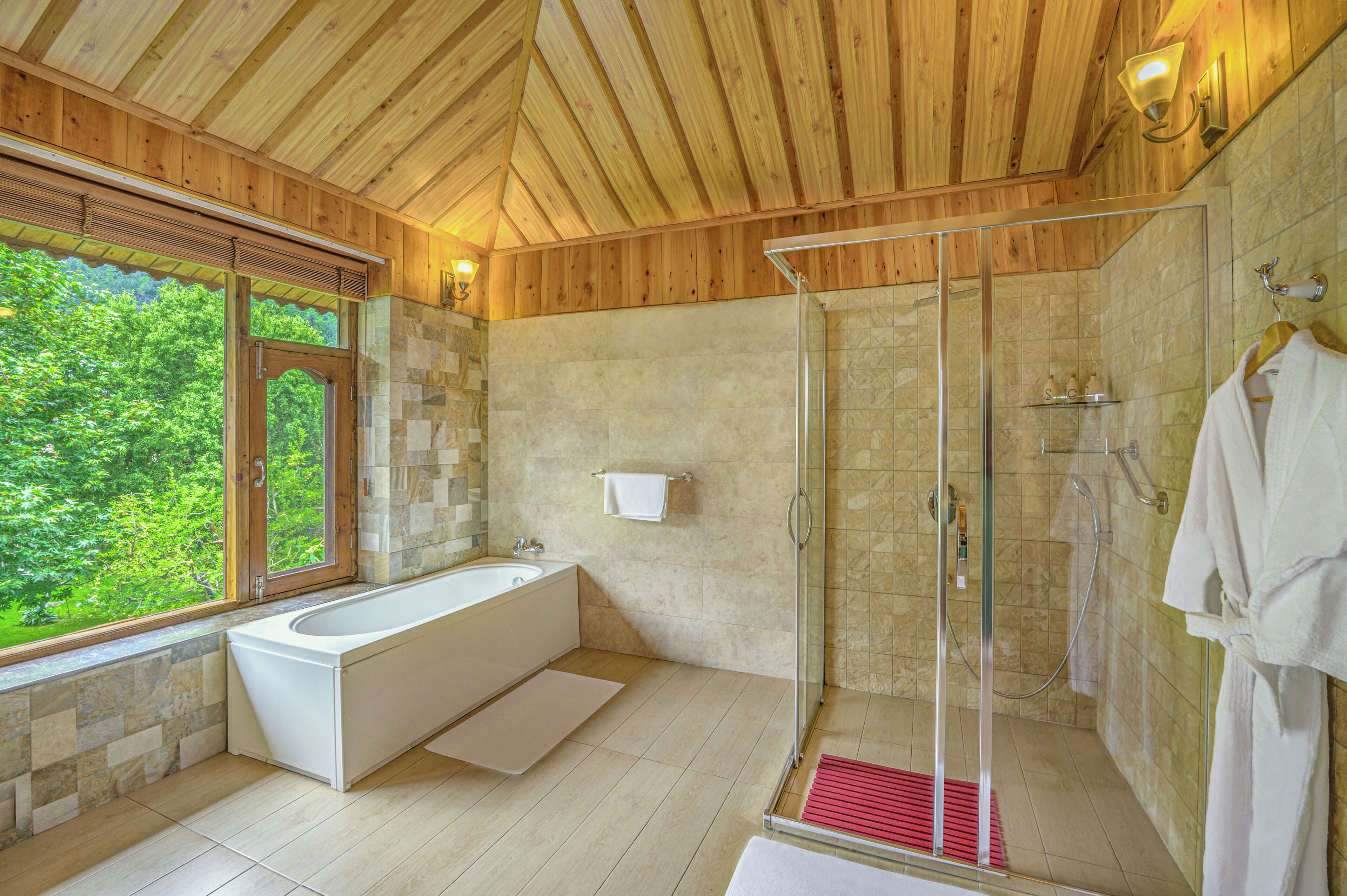 Un baño moderno con ducha de vidrio, bañera independiente y un amplio ventanal con vistas a la vegetación.