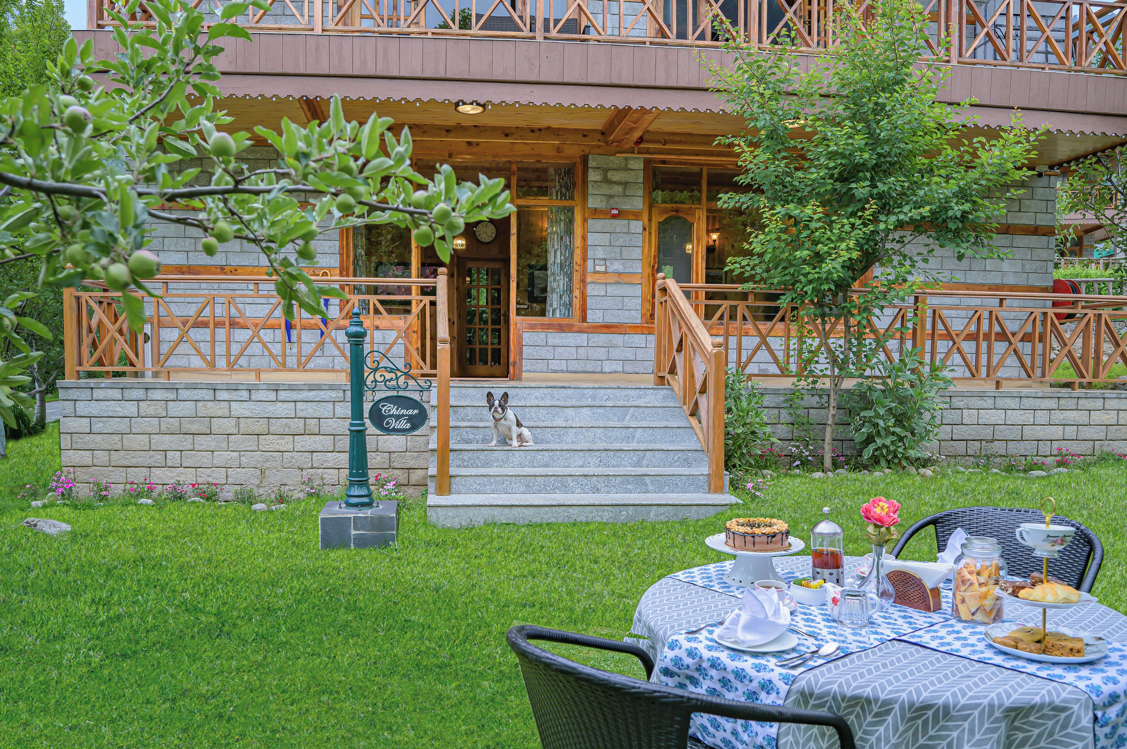 Un bâtiment en bois à deux étages, doté d’une façade en pierre, avec un chien assis sur les marches et une table dressée pour un repas en plein air.