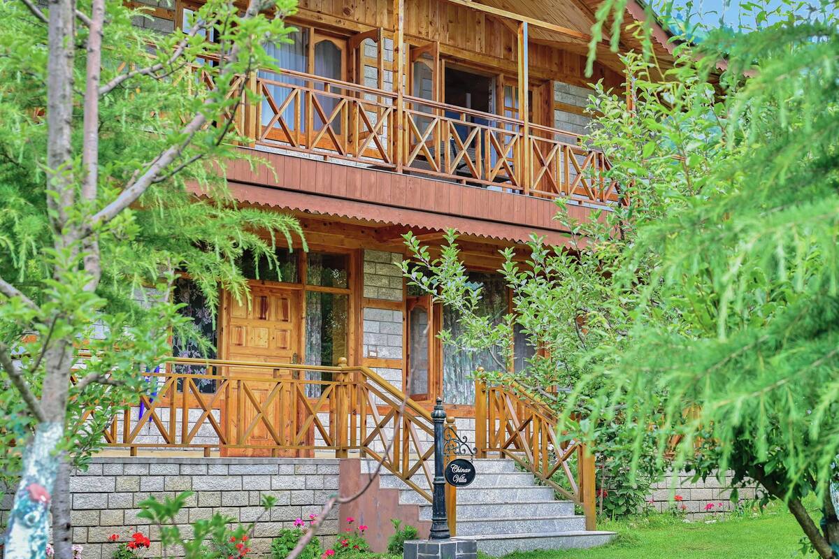 A two-story wooden cabin with a balcony, stone foundation, and a sign reading "Chinar Villa".