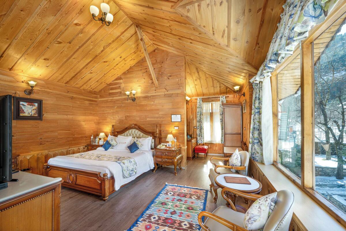 A wooden cabin bedroom with a large bed, bedside tables, a desk, and a window with a view of trees.