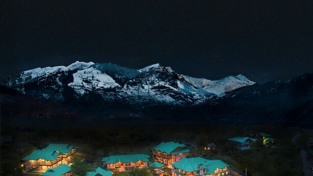 Un refugio de montaña con varias construcciones, cada una con techo verde, frente a un paisaje de picos nevados bajo un cielo estrellado.