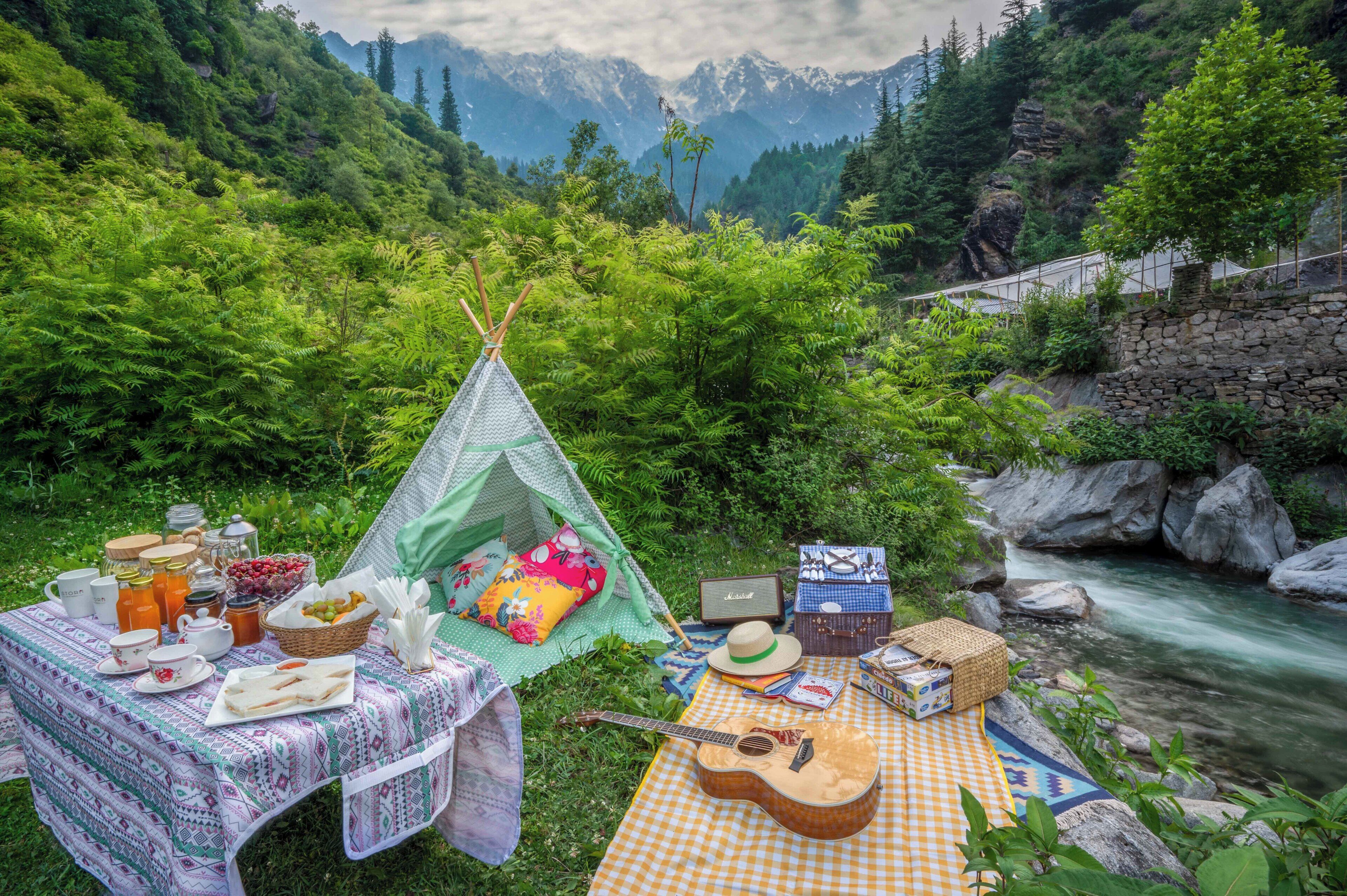 Um piquenique montado com uma tenda, mesa com comida e bebidas, um violão e vista para um rio e montanhas.