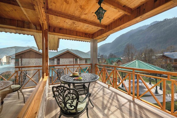 A wooden balcony with a round table and chairs, a view of houses and mountains in the background.