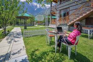 A person sitting at an outdoor table with a laptop, surrounded by a white picket fence, a gazebo, and a building with a stone facade and wooden staircase.