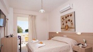 A hotel room with a bed, bedside table, and a desk with a chair. There is a window with curtains, a painting on the wall, and an air conditioning unit. - Akti Corali (Malevizi)