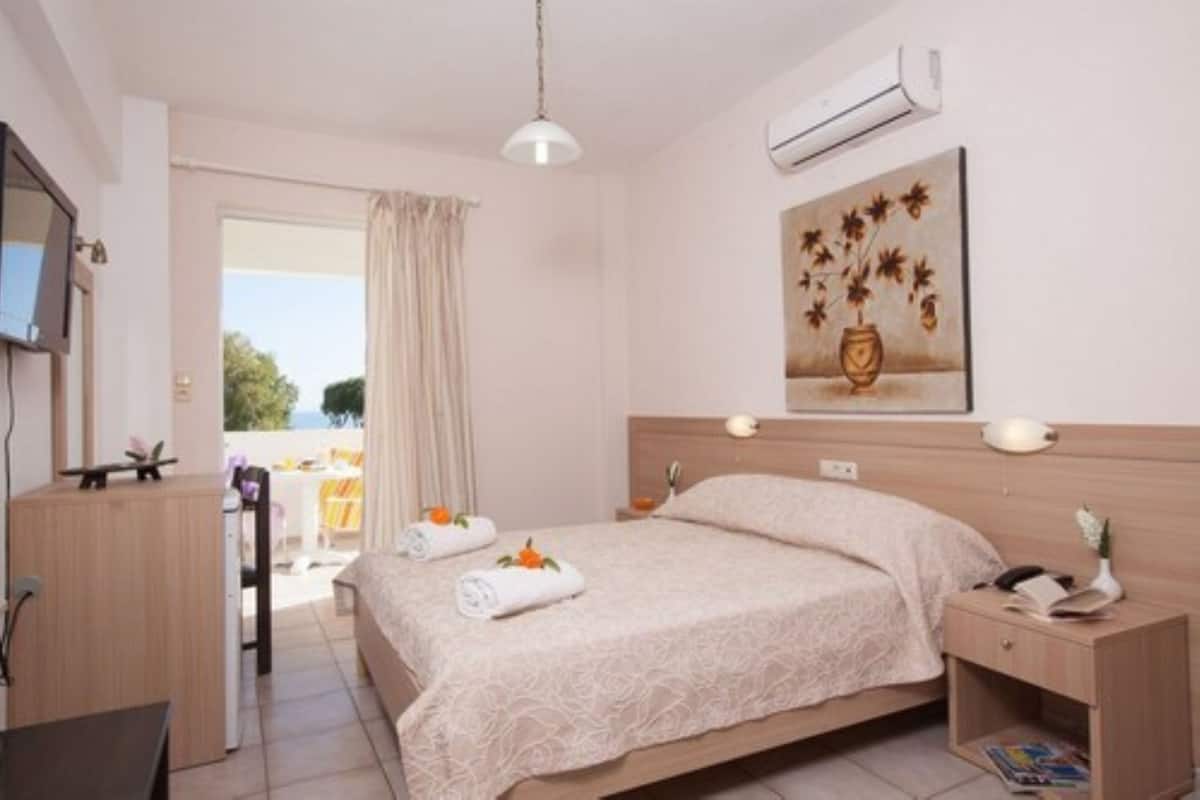 A hotel room with a bed, bedside table, and a desk with a chair. There is a window with curtains, a painting on the wall, and an air conditioning unit.