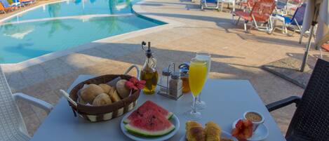 A poolside table with a basket of bread, a plate of watermelon, croissants, and a glass of orange juice.