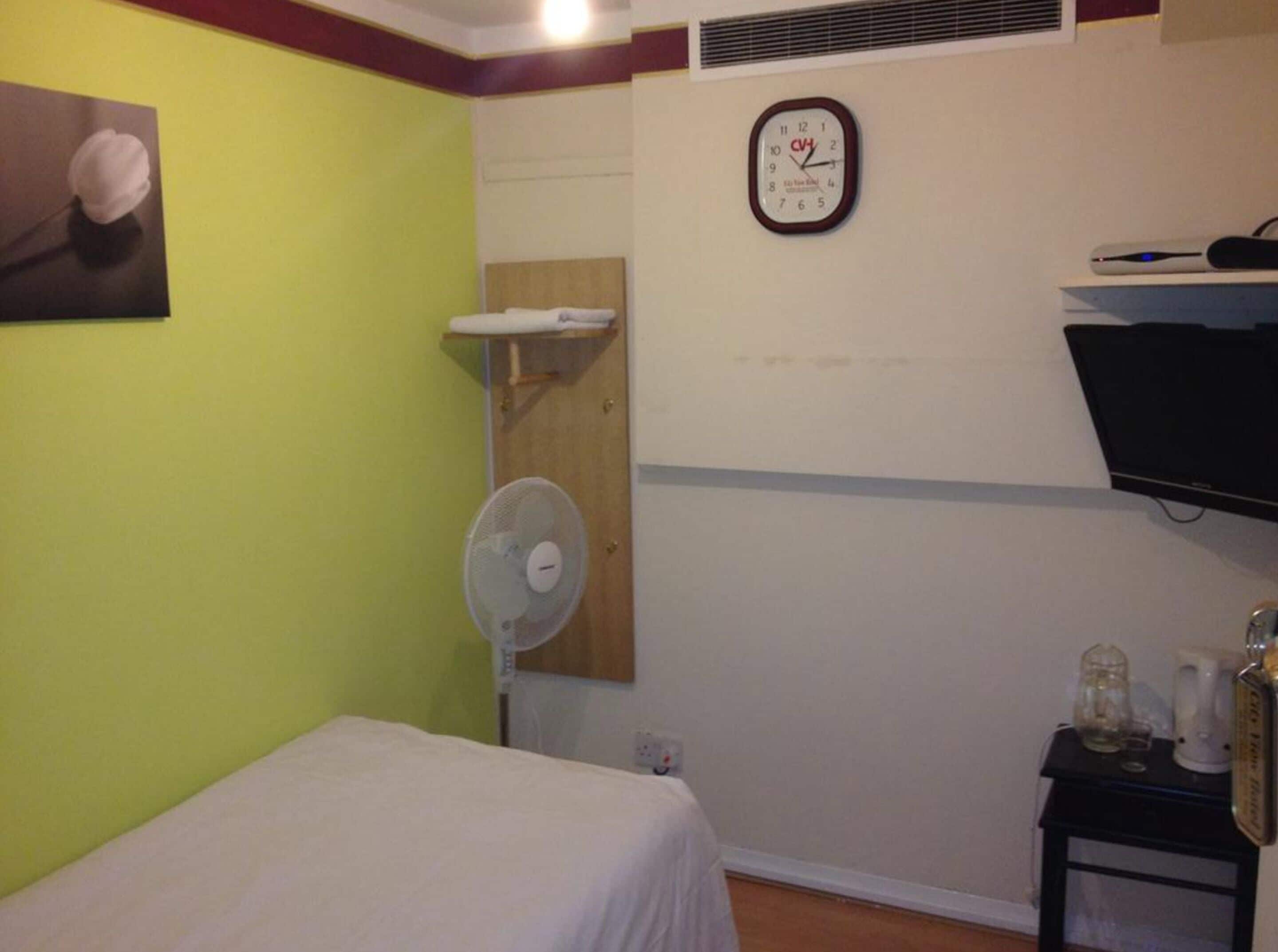 A hotel room with a bed, a wall-mounted clock, a fan, and a small table with a bottle and glasses.