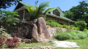 A house with a stone retaining wall, a large rock, and lush greenery.