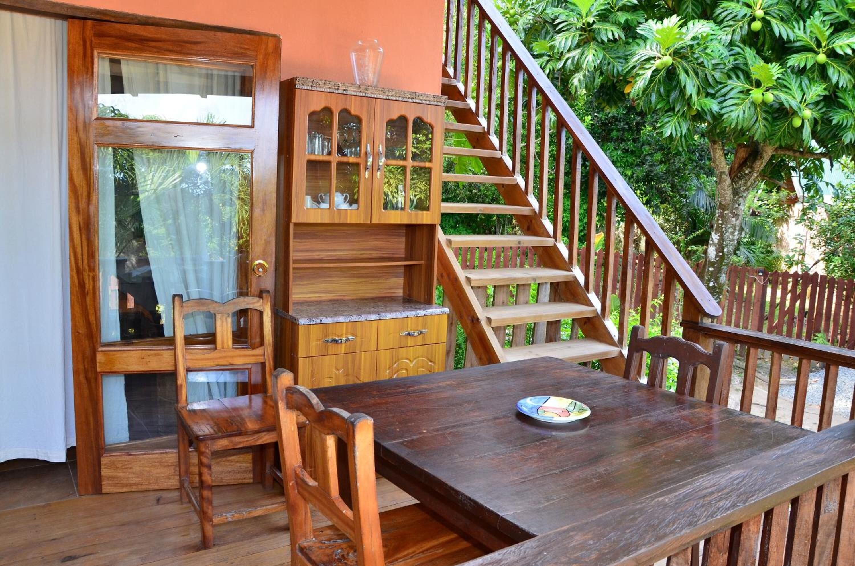 Uma mesa de jantar de madeira com cadeiras, um armário de madeira com portas de vidro e uma escadaria que leva a uma área externa com vegetação.
