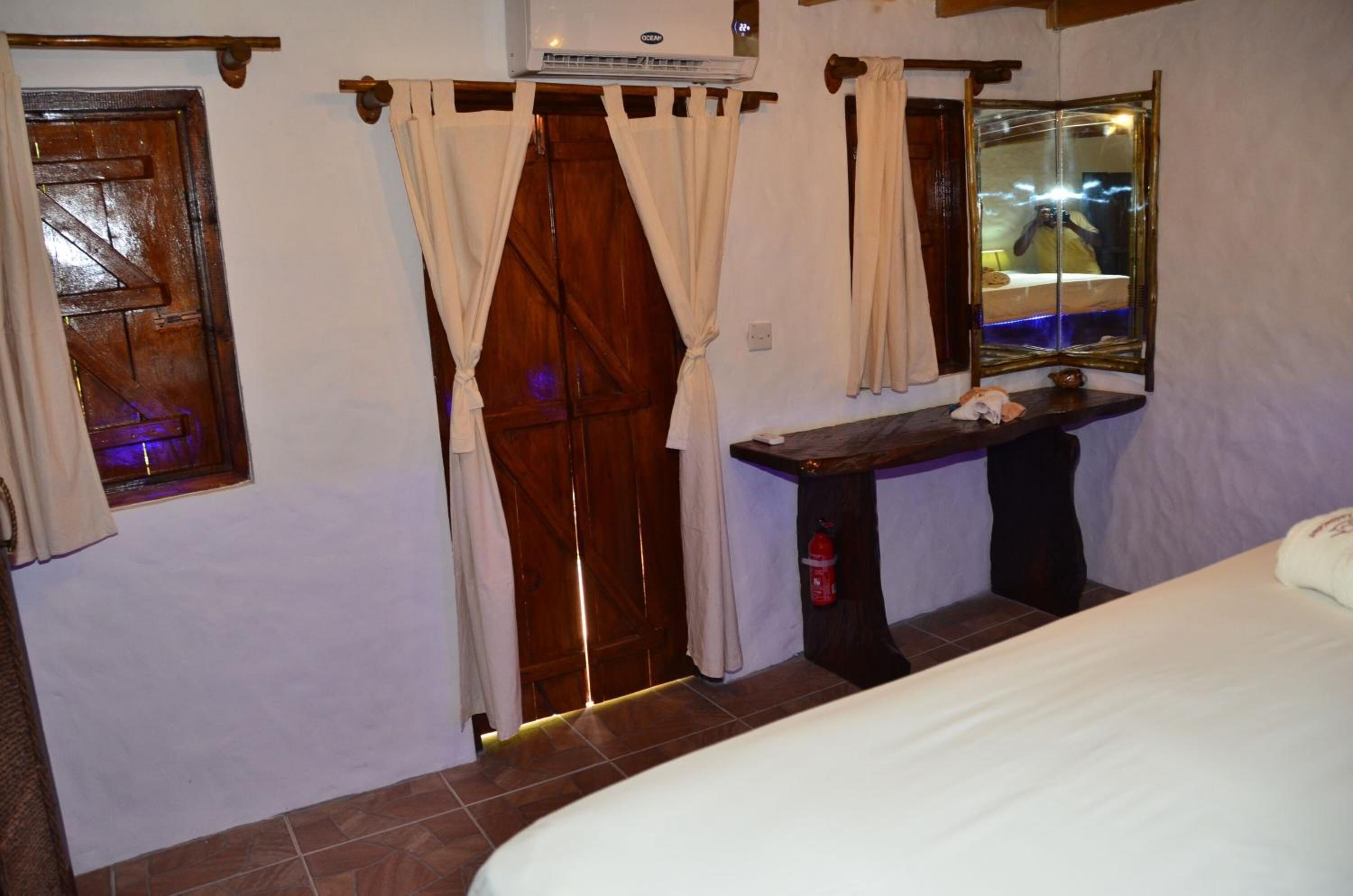 A bedroom with a wooden door, a bed, a bedside table, a mirror, and a window with curtains.