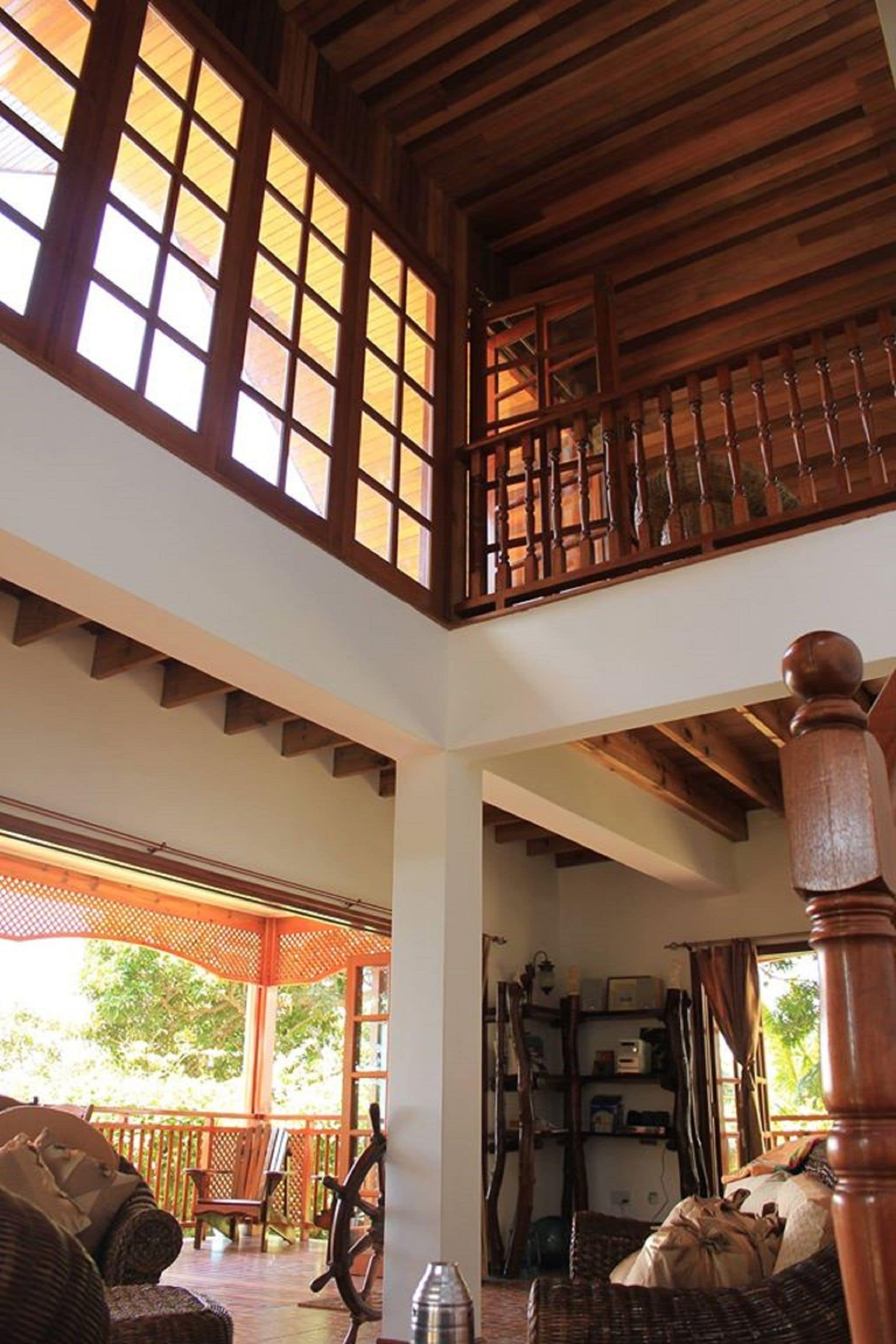 Il s’agit d’une pièce spacieuse avec un plafond haut, des poutres en bois et de grandes fenêtres. Un escalier en bois mène à un niveau supérieur. La pièce dispose d’un coin salon confortable, avec des meubles en osier et une table en bois.