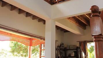 Il s’agit d’une pièce spacieuse avec un plafond haut, des poutres en bois et de grandes fenêtres. Un escalier en bois mène à un niveau supérieur. La pièce dispose d’un coin salon confortable, avec des meubles en osier et une table en bois.