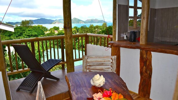 A wooden deck with a table set for a meal, overlooking a scenic view of mountains and the sea. - Kaz Ladouceur (La Digue)