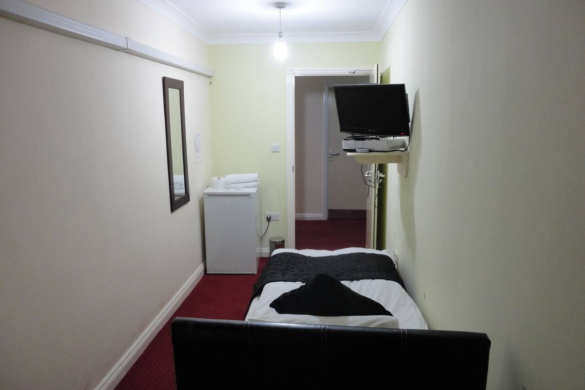 Habitación de hotel estrecha con una cama, un refrigerador blanco pequeño, un televisor montado en la pared y una puerta que da a otra habitación.
