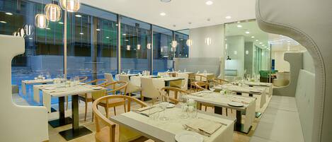 A modern restaurant with multiple dining tables, chairs, and pendant lighting.
