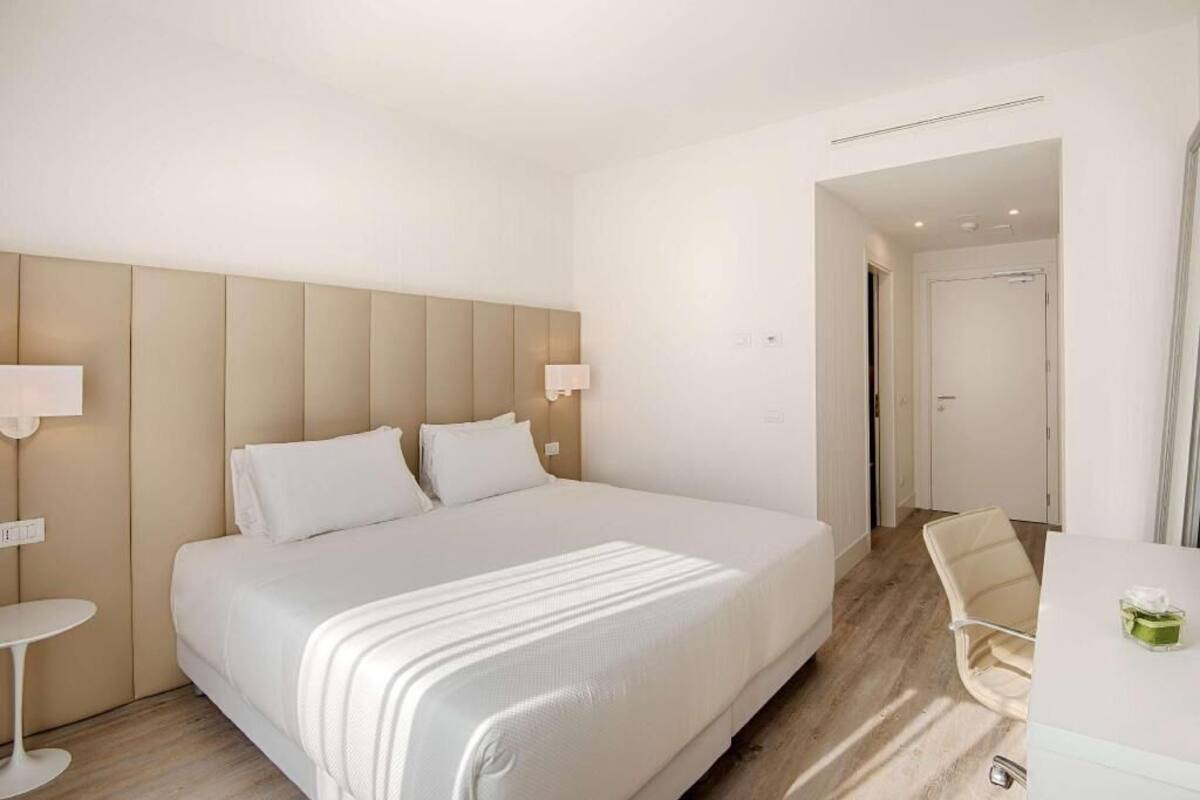 A hotel room with a large bed, a bedside table, a chair, and a door leading to another room.