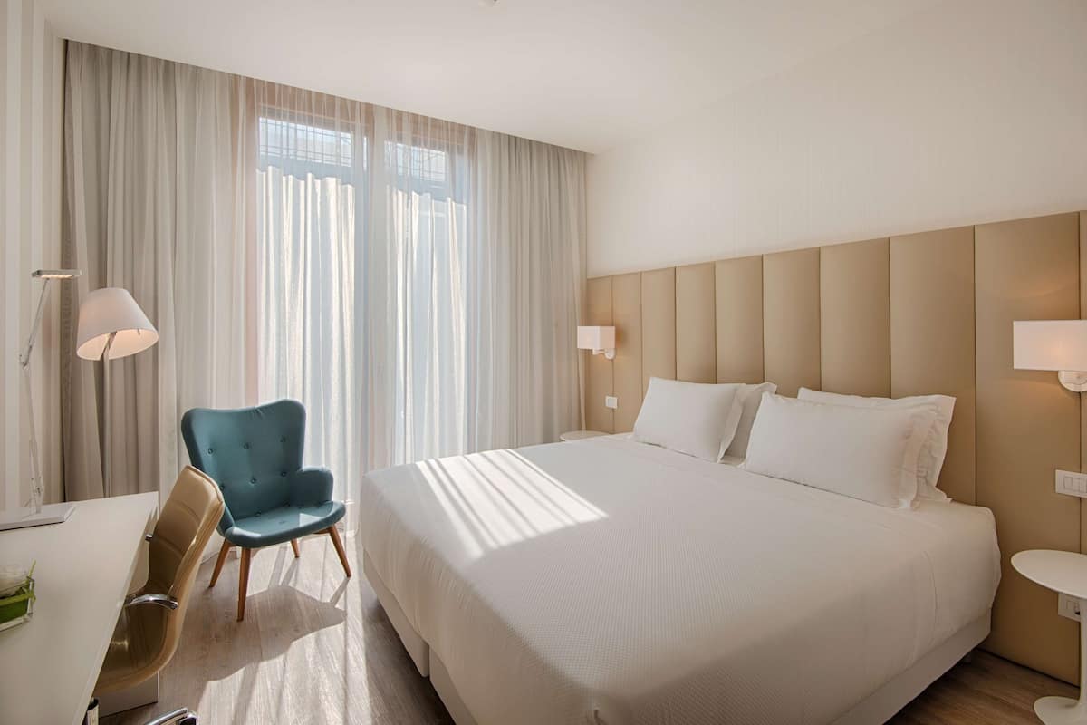 A hotel room with a large bed, a desk, a chair, and a window with sheer curtains.