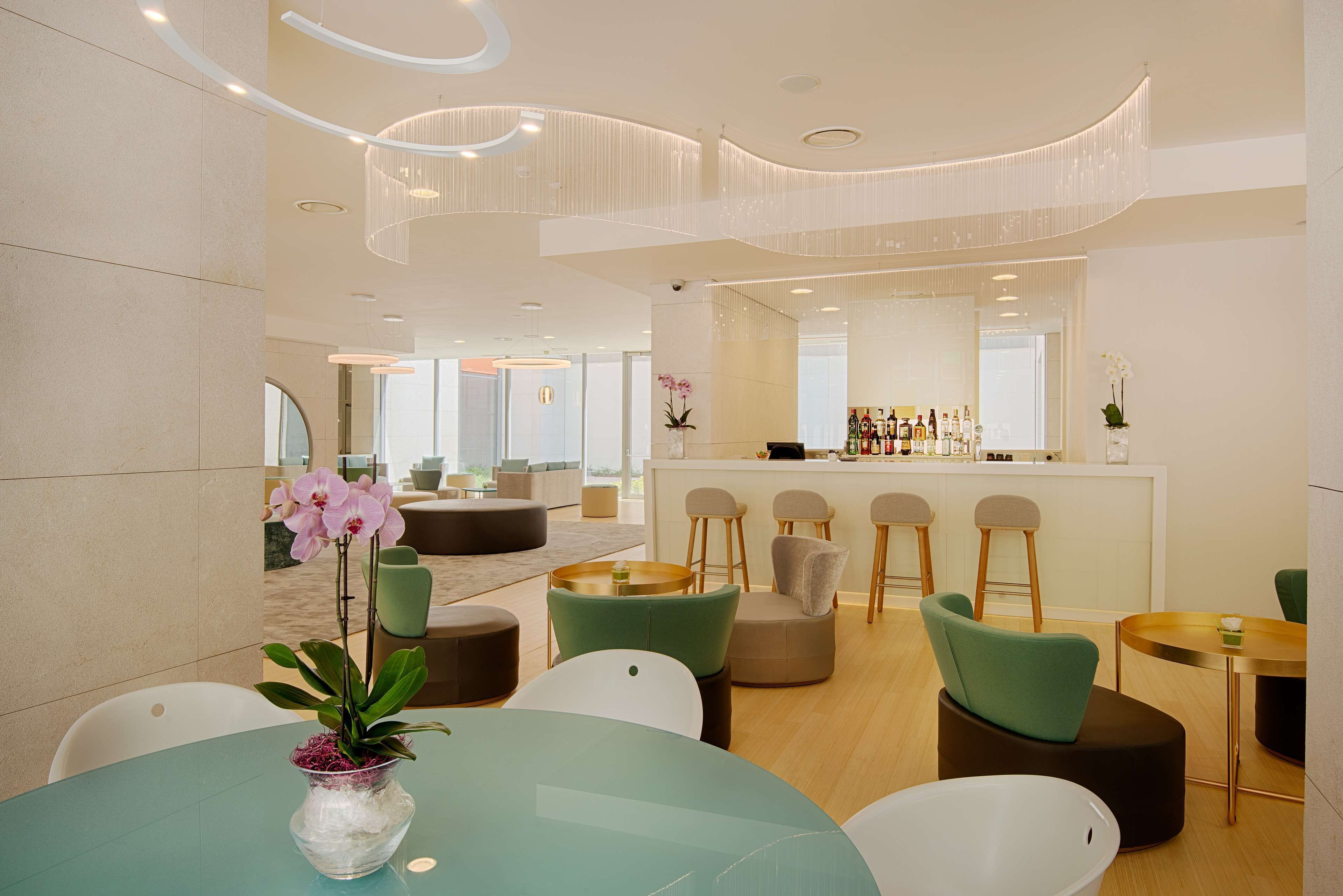 A modern hotel lobby with round tables, green and white chairs, a bar area, and a large mirror reflecting the space.