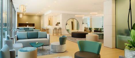 A modern hotel lobby with a seating area, a reception desk, and a kitchen in the background.