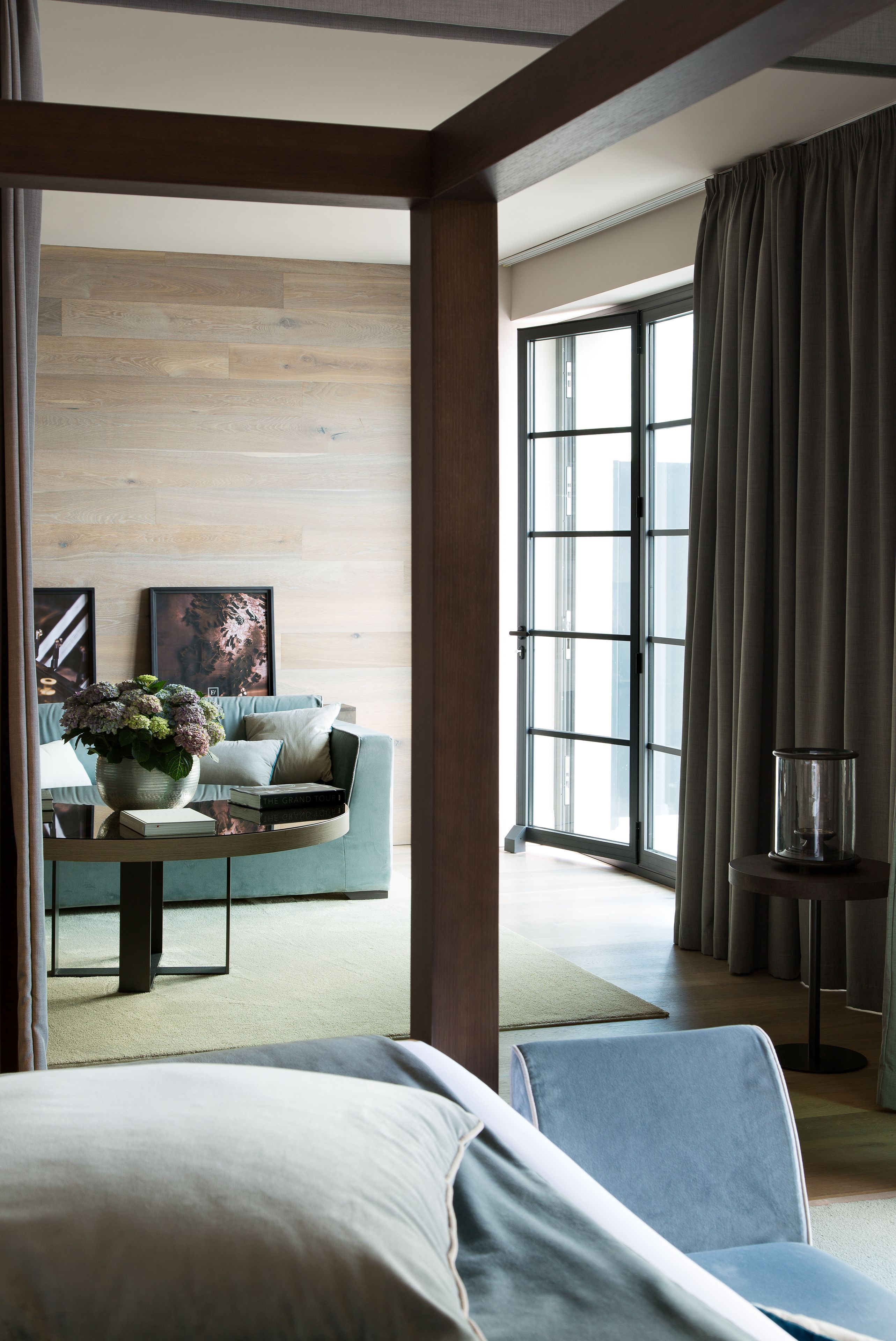 A modern hotel room with a large window, a sofa, a coffee table with a vase of flowers, and a bed.