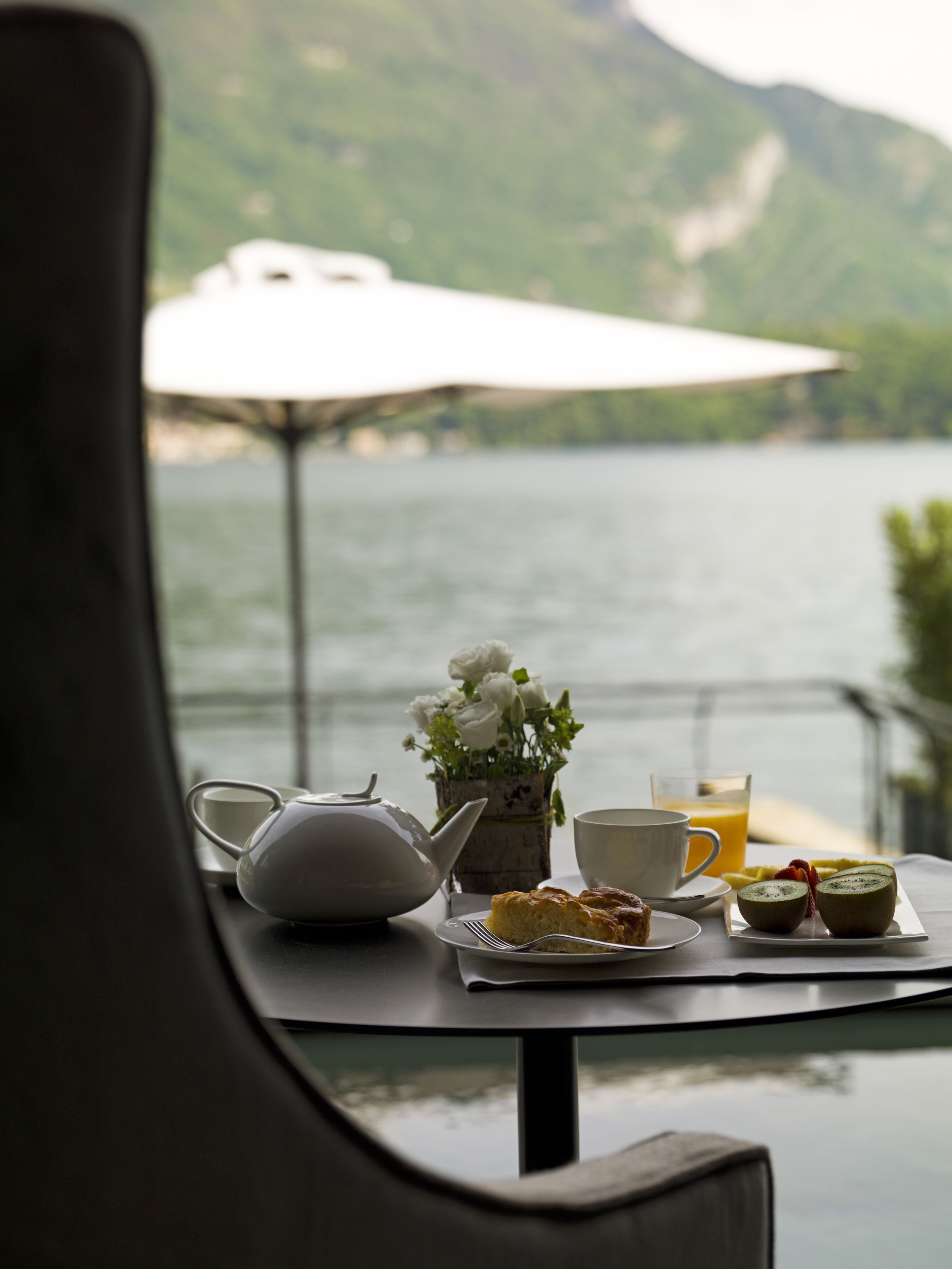 A table with a teapot, a cup, a glass of orange juice, and a plate with food, set against a scenic lake view.