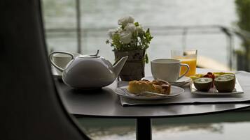 A table with a teapot, a cup, a glass of orange juice, and a plate with food, set against a scenic lake view.
