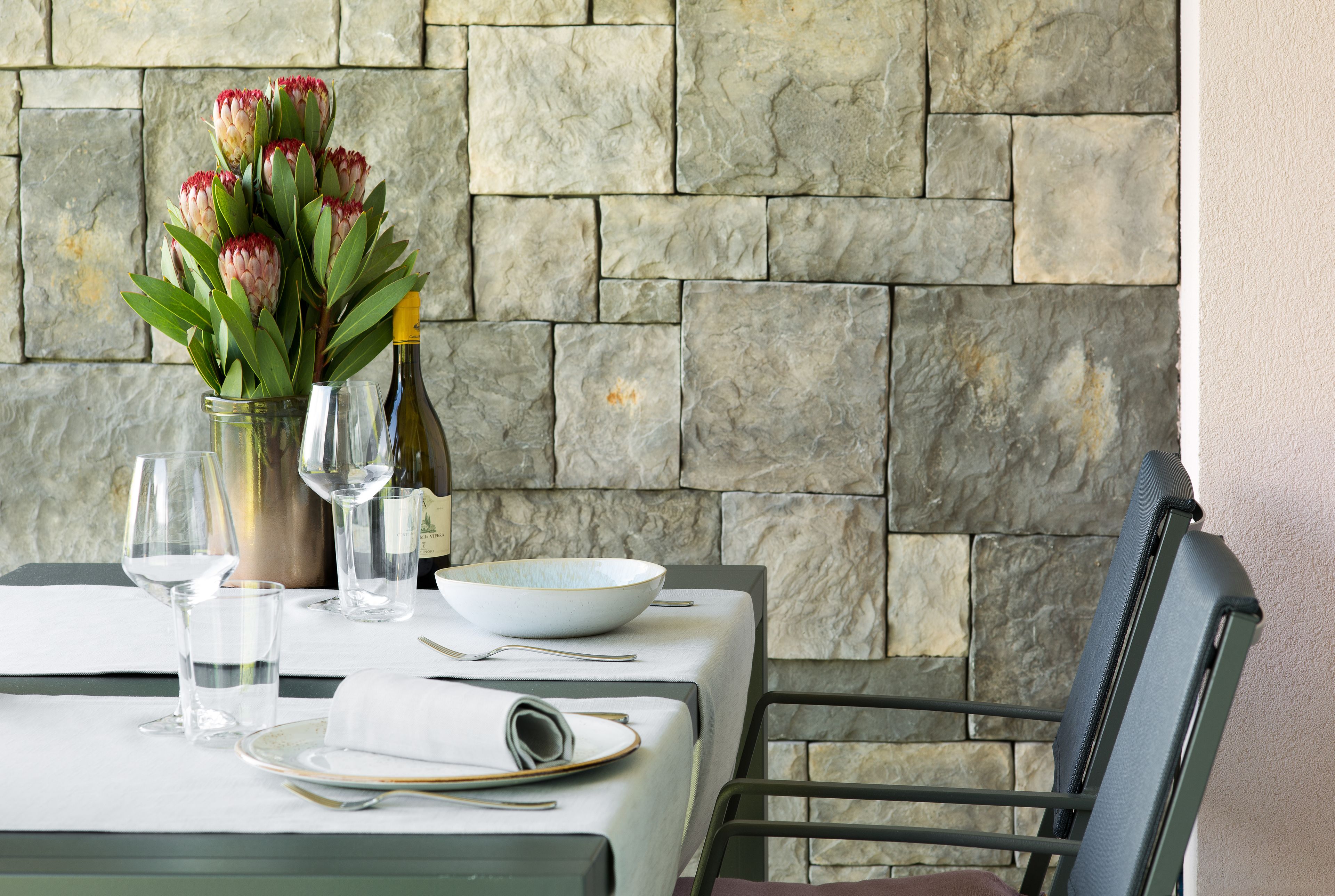 Mesa servida con un jarrón de flores, copas de vino y platos, con un fondo de pared de piedra.