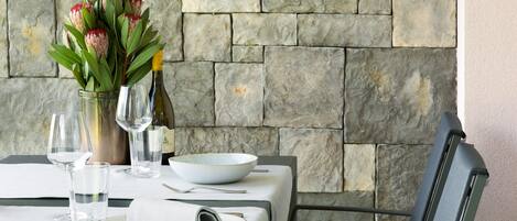 A table set with a vase of flowers, wine glasses, and plates, against a stone wall backdrop.