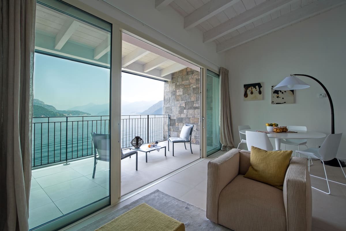Een moderne woonkamer met een bank, een tafel en stoelen. Via grote glazen deuren is een balkon zichtbaar met uitzicht op zee en bergen.