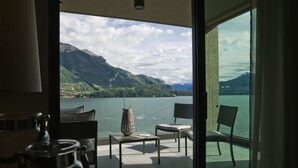 A room with a view of a lake and mountains. There is a table with chairs and a vase.